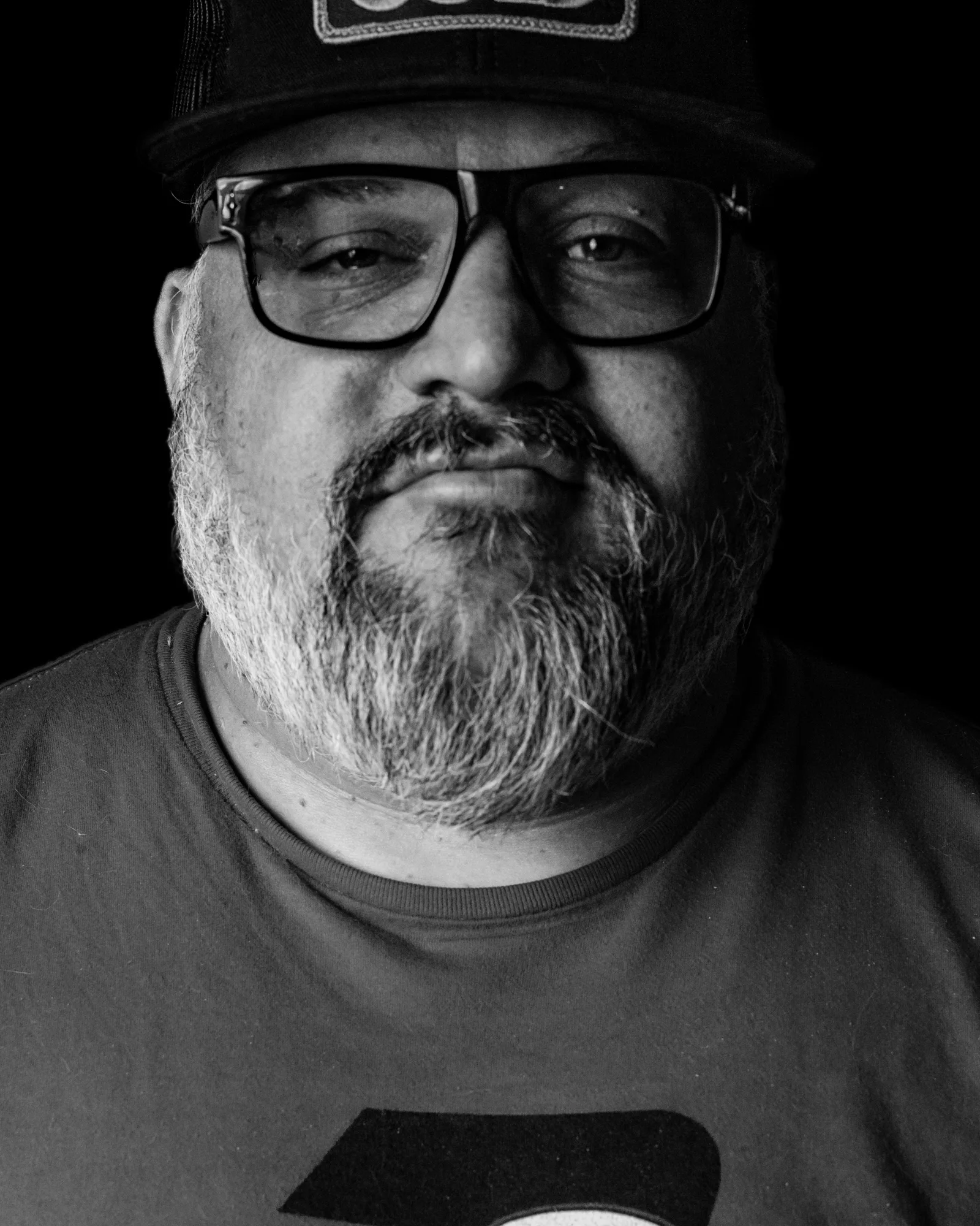 Black-and-white close-up of a bearded man's chin, neck, and dark t-shirt, mouth slightly upturned in a faint smile.