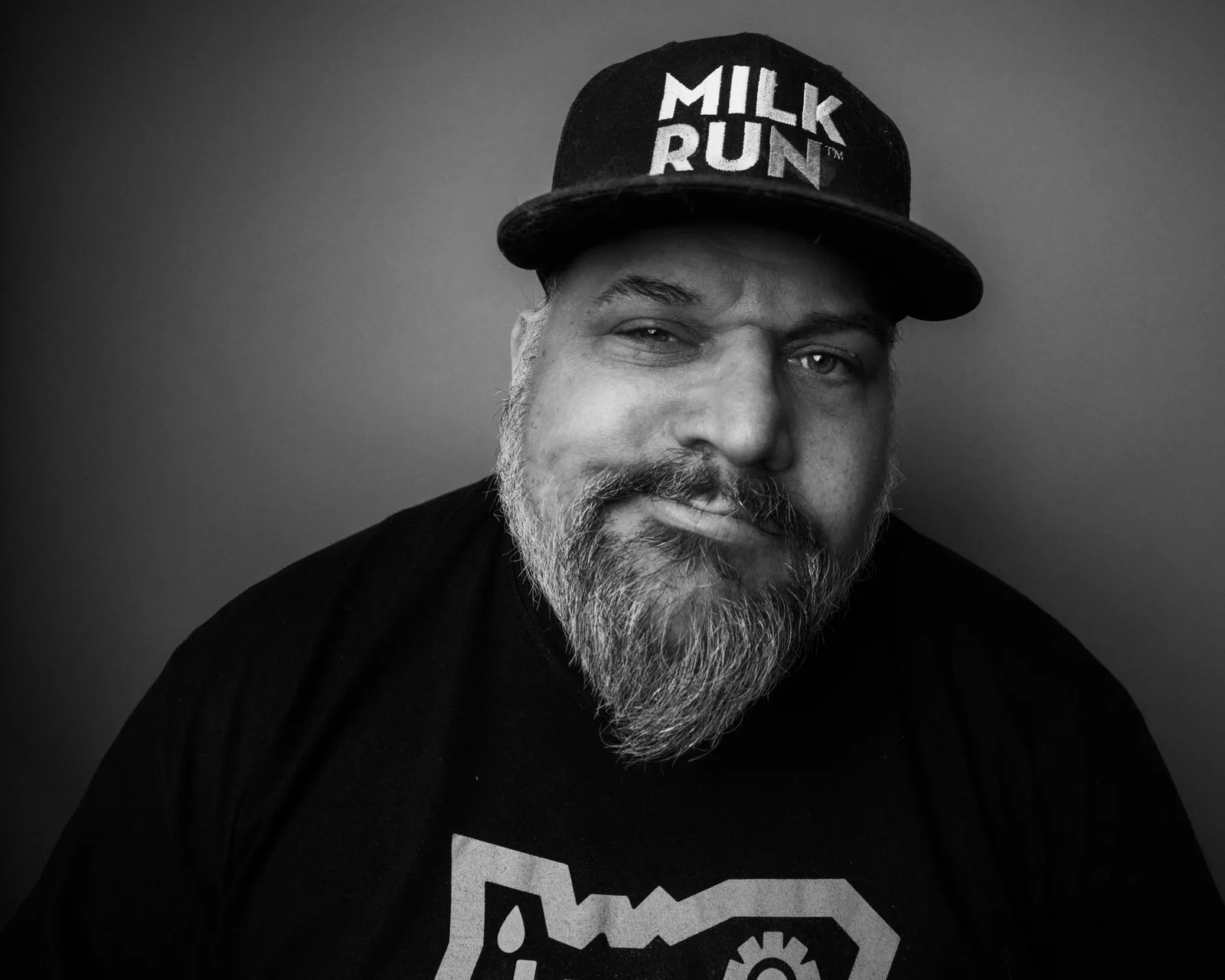 Black-and-white close-up of a bearded man in a snapback cap reading 'RUN', smirking slightly at the camera in a black t-shirt.