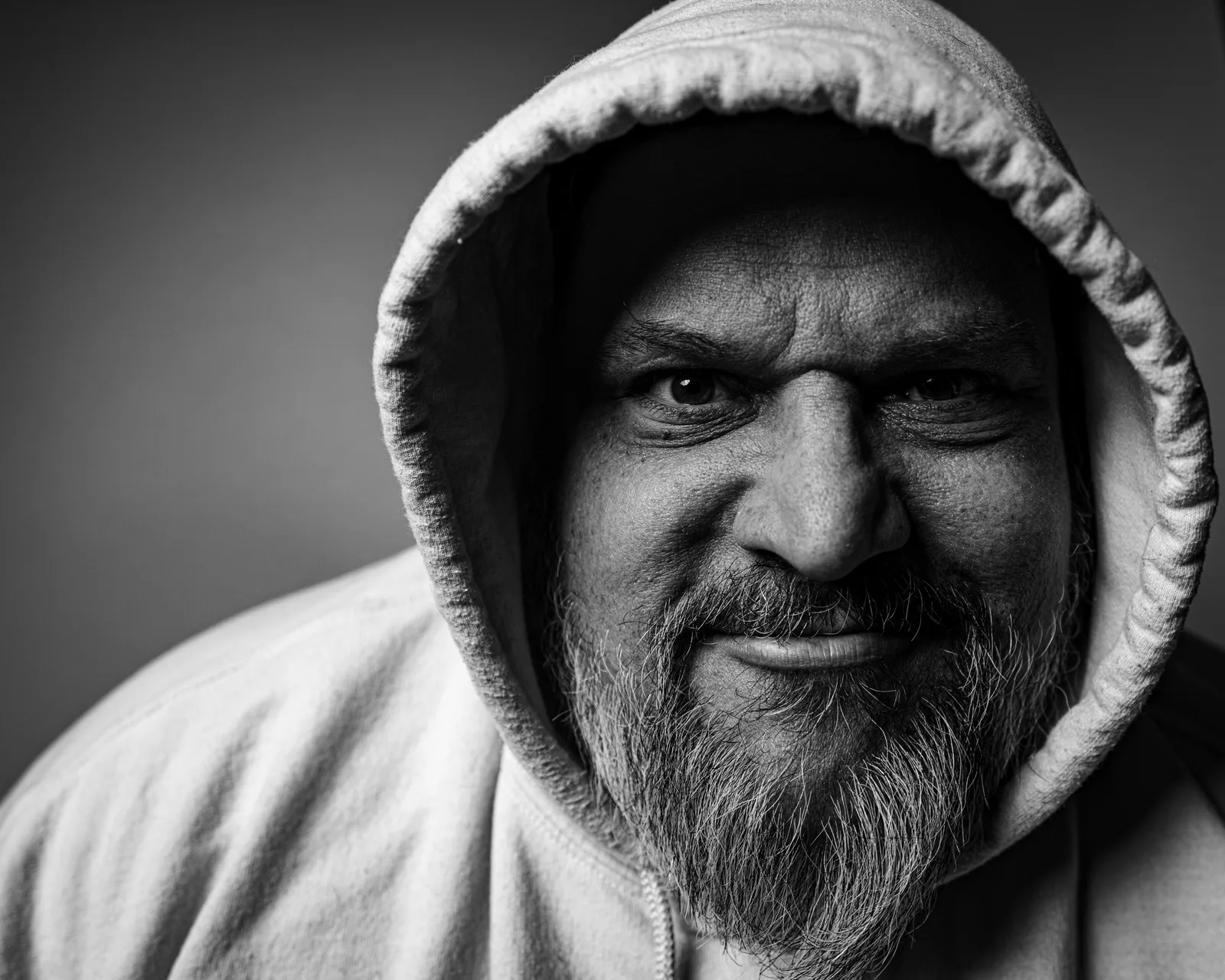 Black-and-white close-up of a bearded man in a hoodie, smirking slightly with a mischievous glint in his eyes.