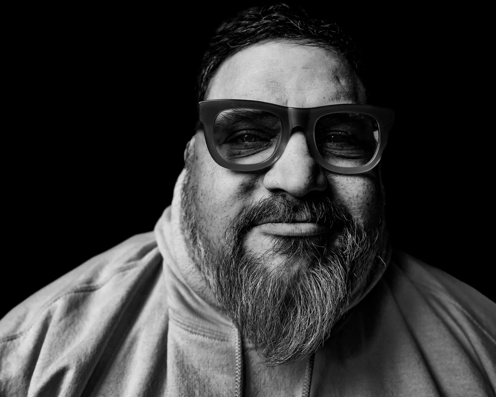 Black-and-white tight portrait of a bearded man in glasses and a high-collared jacket, faint smile, against black.