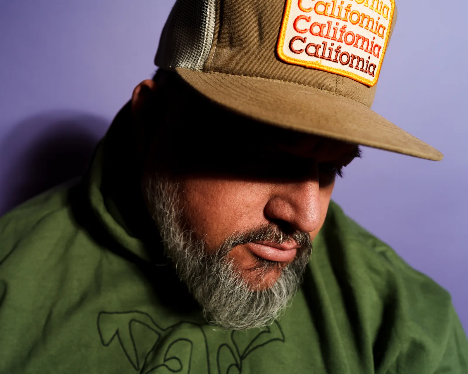 Close-up of a bearded man looking down under a tan trucker cap with a 'California California California' patch, purple background.