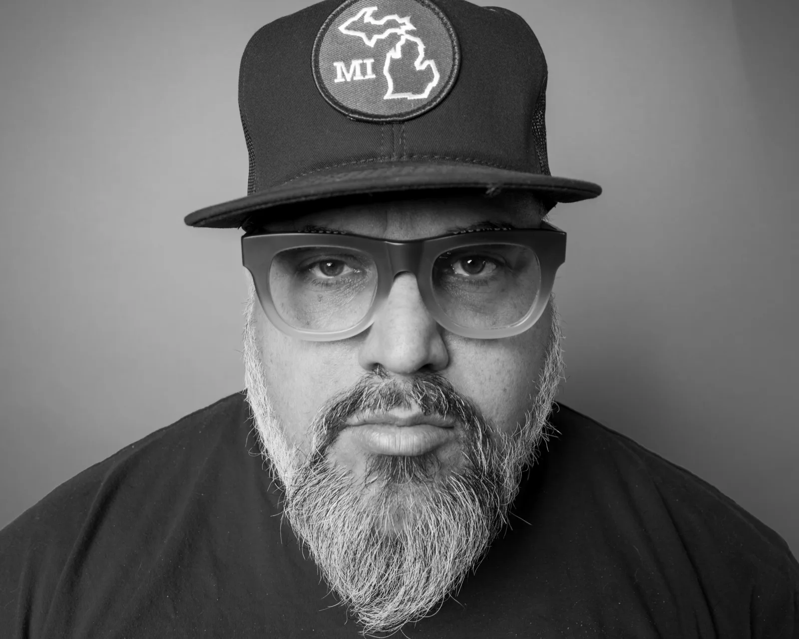 Black-and-white close-up of a bearded man in a fedora and thick-framed glasses, staring directly at the camera.