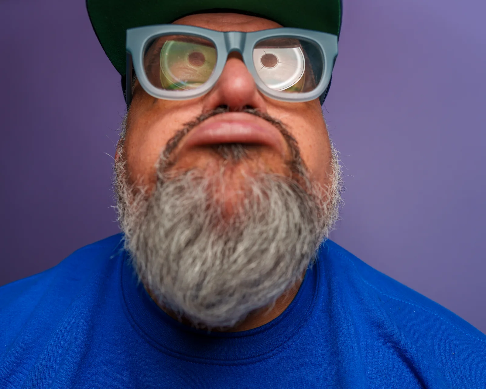Extreme close-up of a bearded man tilting his face up toward the camera, wearing pale blue glasses and a green cap, against purple.