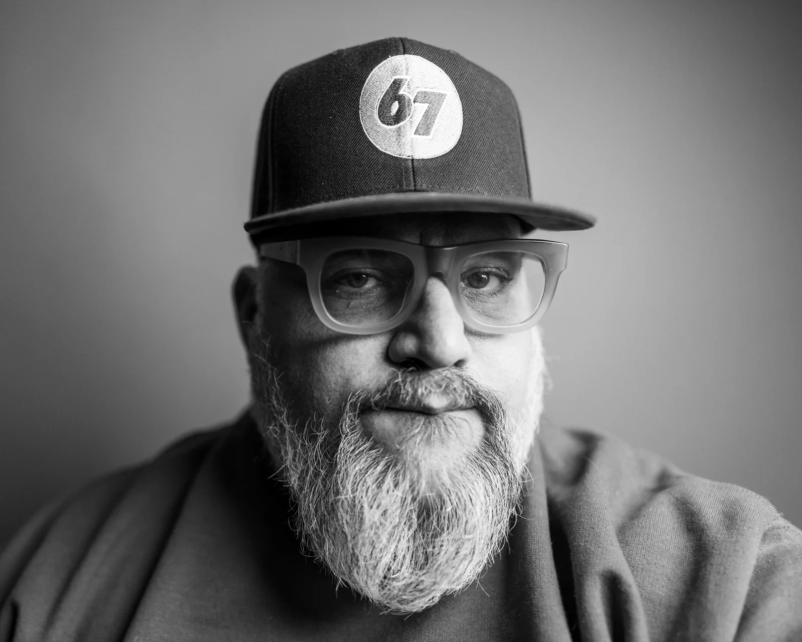 Black-and-white close-up of a bearded man in glasses and a cap, wearing a soft hooded sweatshirt, gazing calmly at the camera.
