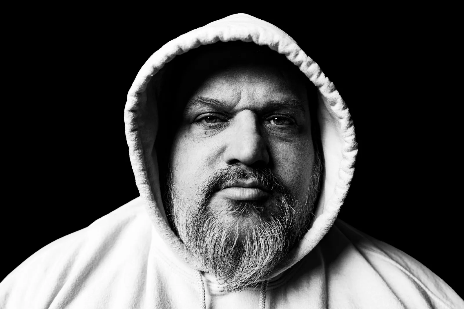 Black-and-white tight portrait of a bearded man in a white hoodie pulled up over his head, gazing directly at the camera against black.