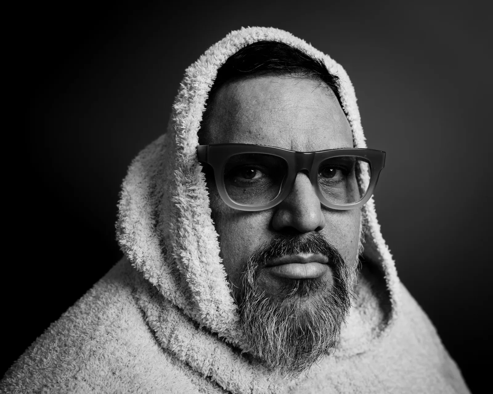 Black-and-white close-up of a bearded man in thick-rimmed glasses and a fuzzy sherpa hood, expression somber and direct.