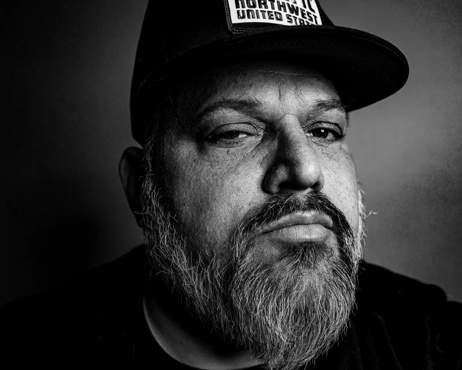 Black-and-white tight close-up of a bearded man in a dark cap, heavy-lidded eyes looking down at the camera, somber expression.