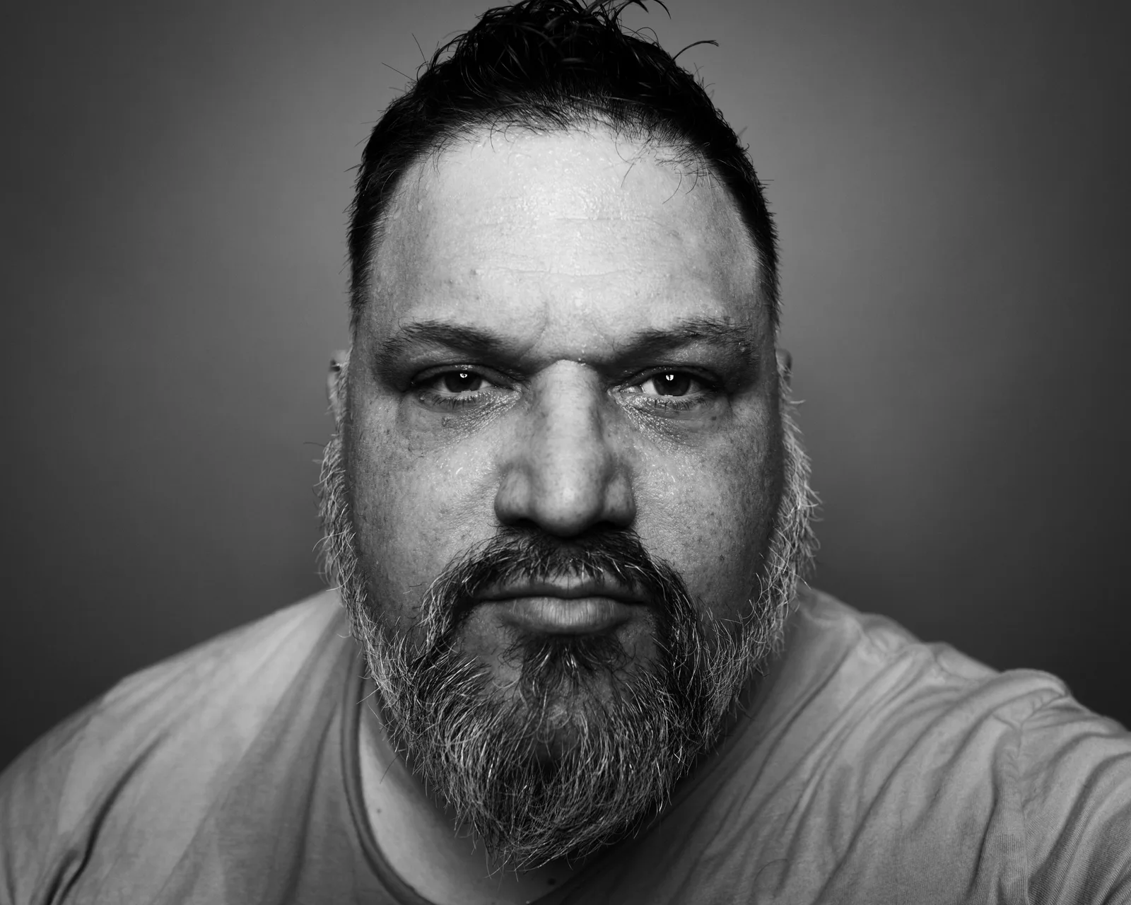Black-and-white tight close-up of a bearded man's face, sweat on his forehead, eyes locked directly on camera.