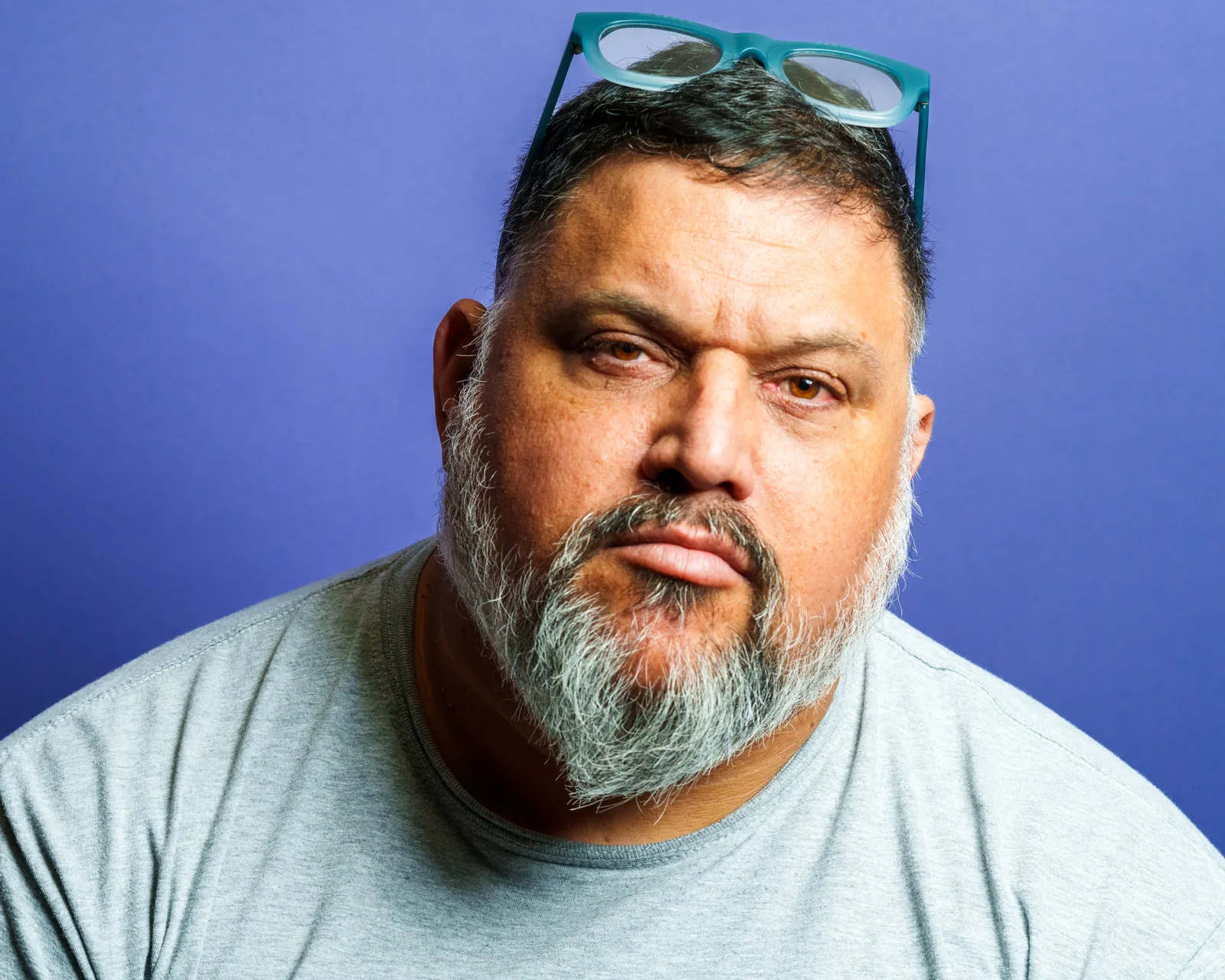 Close-up of a bearded man in a gray t-shirt against a purple background, eyes steady, mouth set in a quiet half-smile.