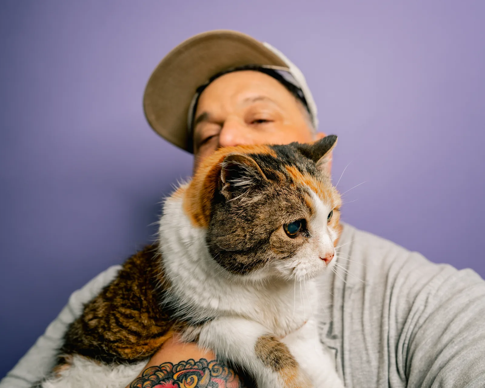 Close-up of a calico cat held up to the face of a man in a tan cap, his eyes closed, smiling against a purple backdrop.