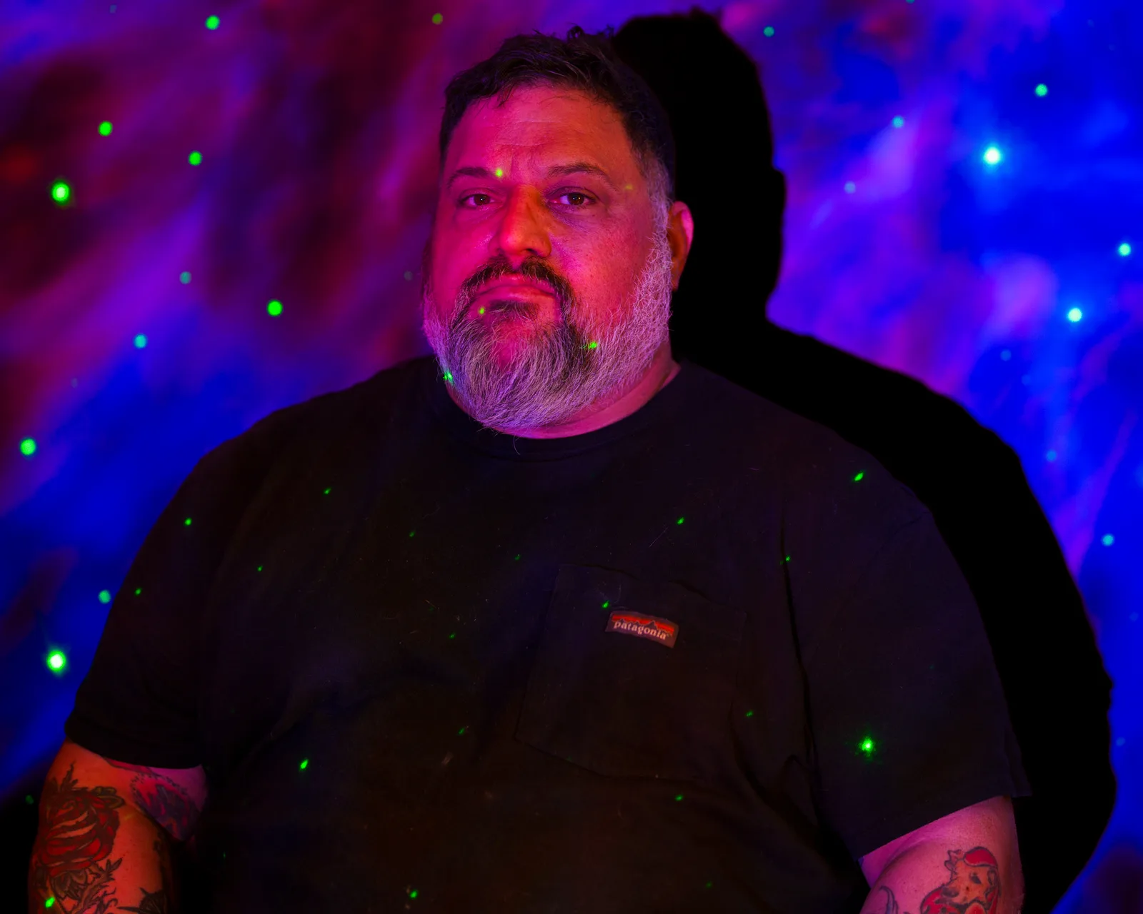 Bearded man in black t-shirt lit by pink and blue light, green star-like dots projected across him and the wall behind.