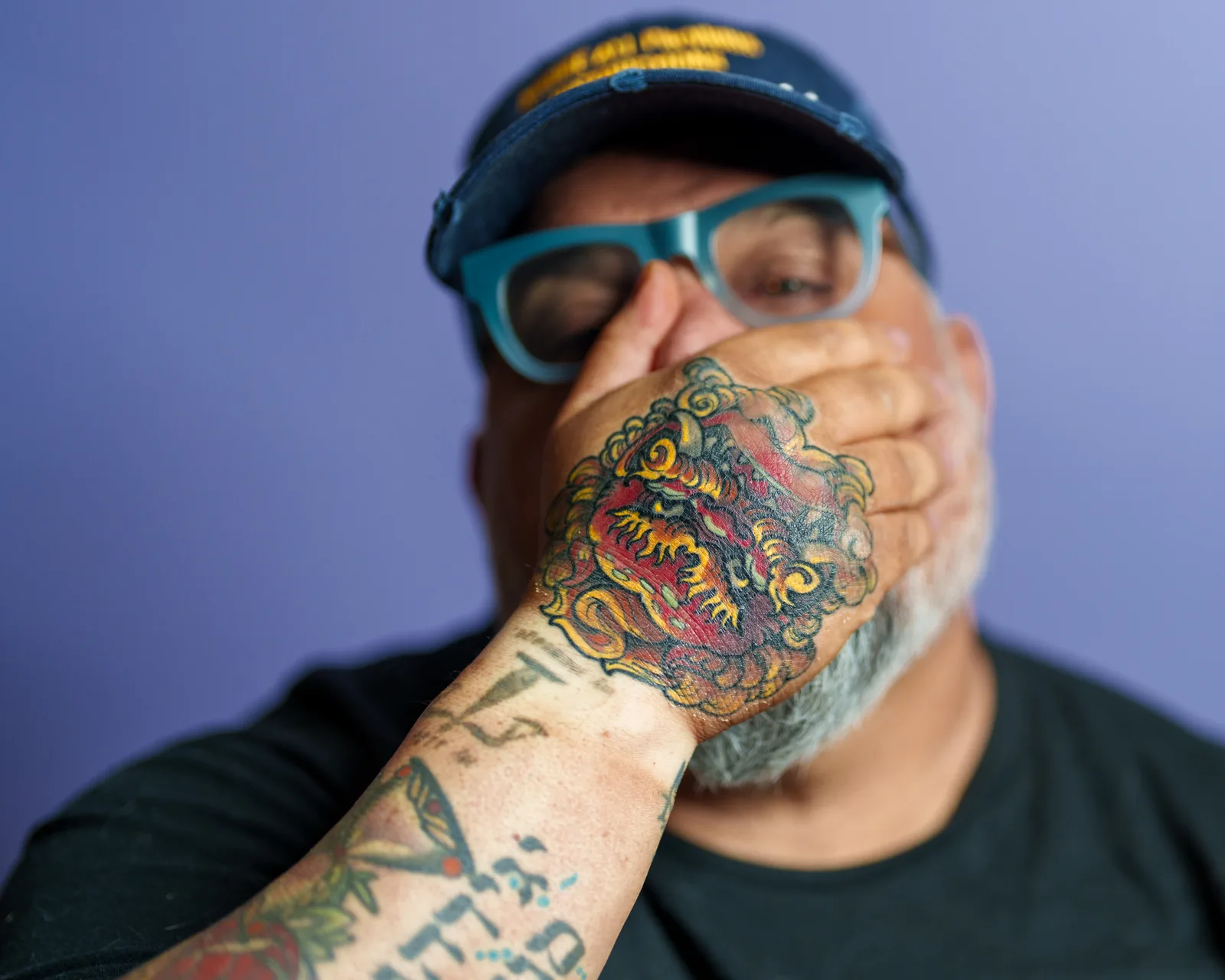 Bearded man's face partly hidden behind his tattooed hand, showing a vivid red and yellow foo dog tattoo and inked forearm.