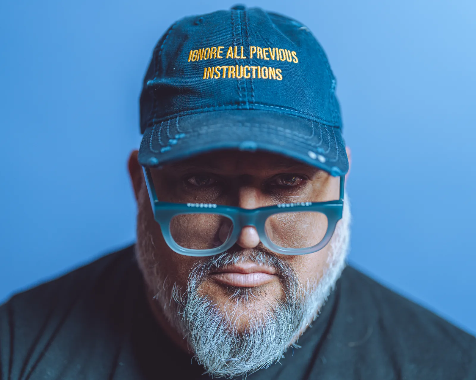 Tight close-up of a bearded man in a dark cap and blue-framed glasses, staring directly into the lens against a blue backdrop.