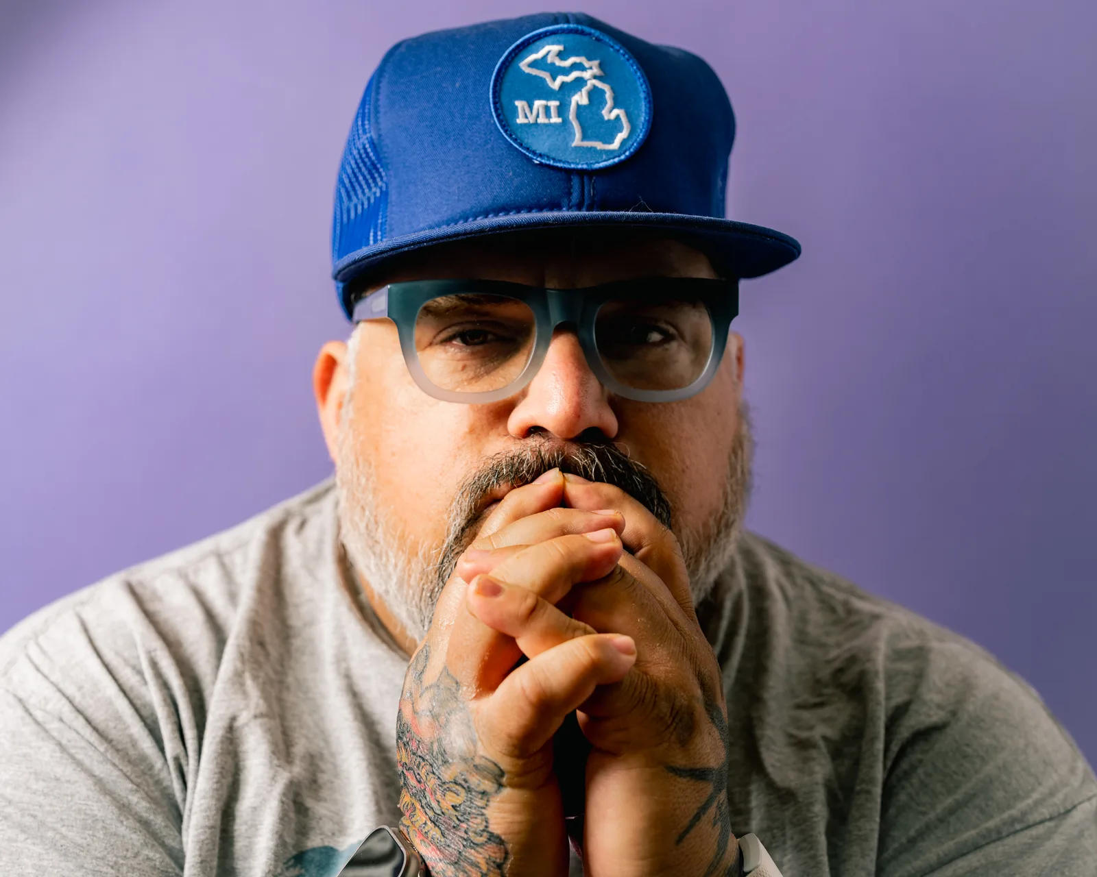 Close-up of a bearded man in a gray t-shirt, hands clasped at his mouth, blue glasses pushed up on his forehead, against purple.