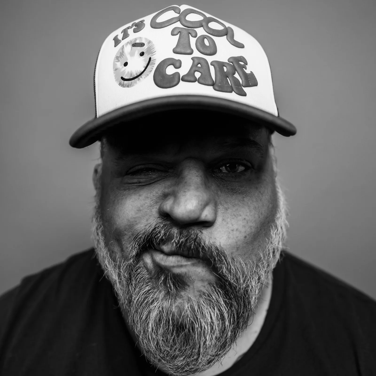 Black-and-white tight crop showing eyes and white trucker hat reading 'IT'S COOL TO CARE' with tie-dye smiley face.