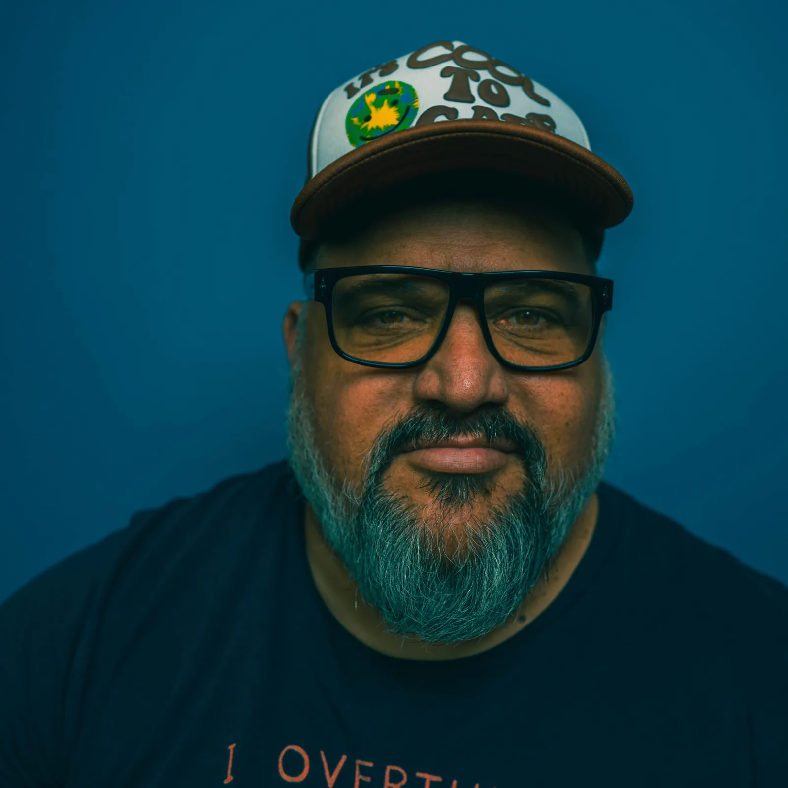 Tight crop on a bearded man's face wearing black square glasses and a brown and white trucker hat reading 'It's Cool To Care' against a teal wall.