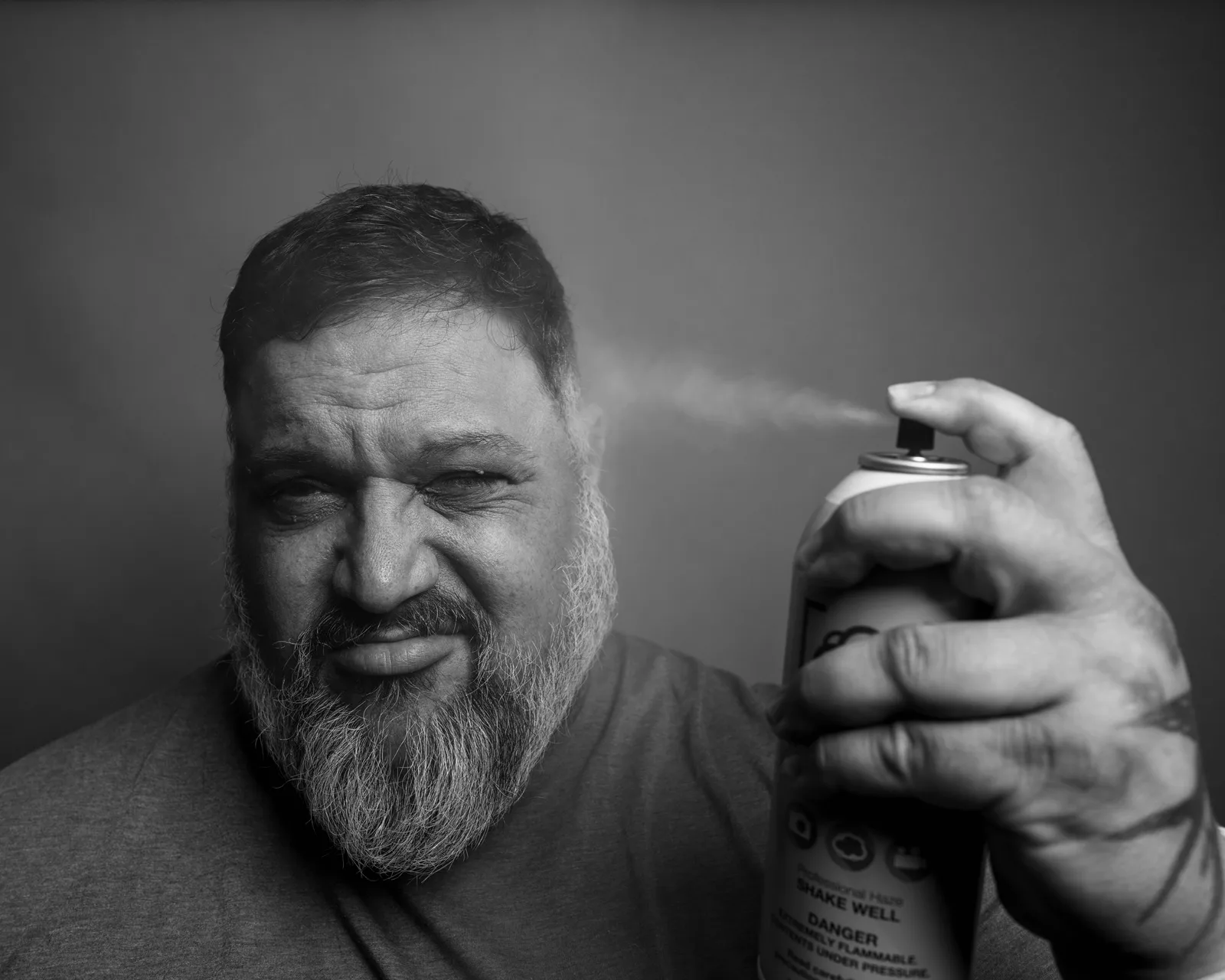 Black-and-white close-up of a bearded man wincing as he sprays an aerosol can directly toward his own face.