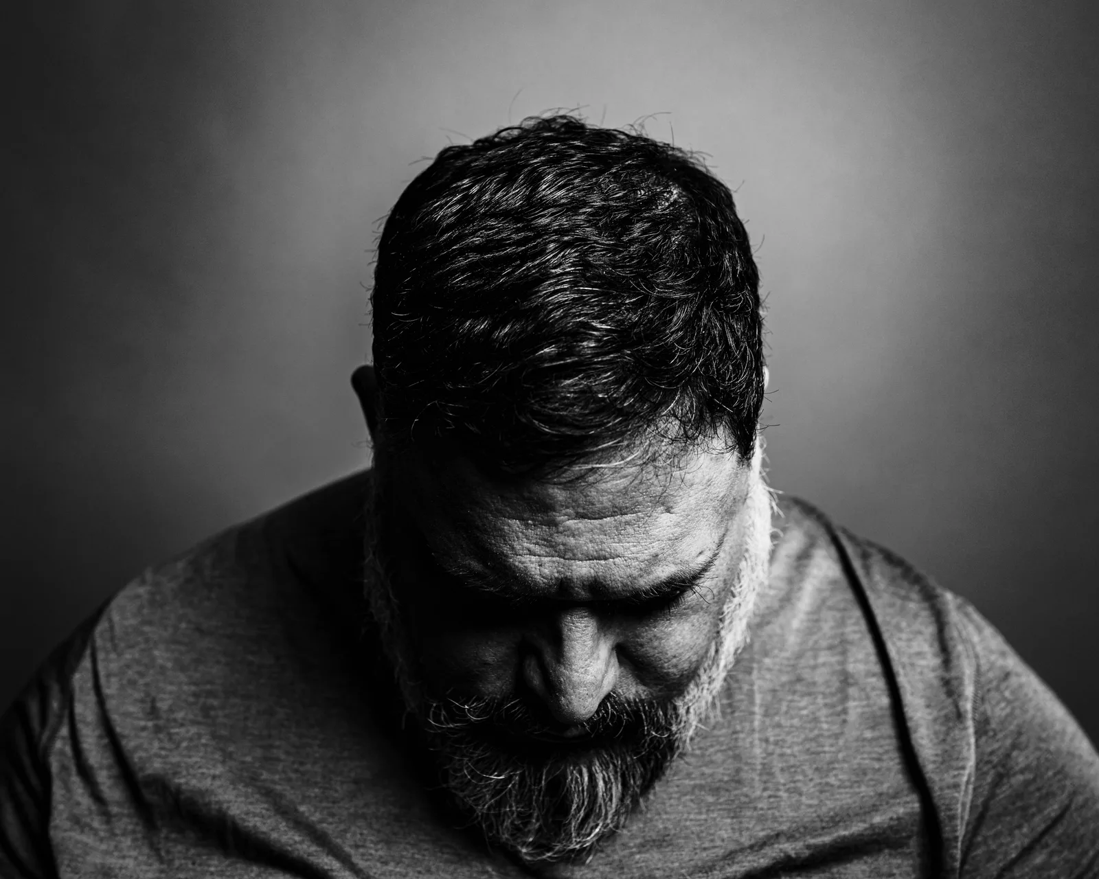 Black-and-white close-up of a bearded man with head bowed, eyes downcast, forehead lined, wearing a soft gray t-shirt.