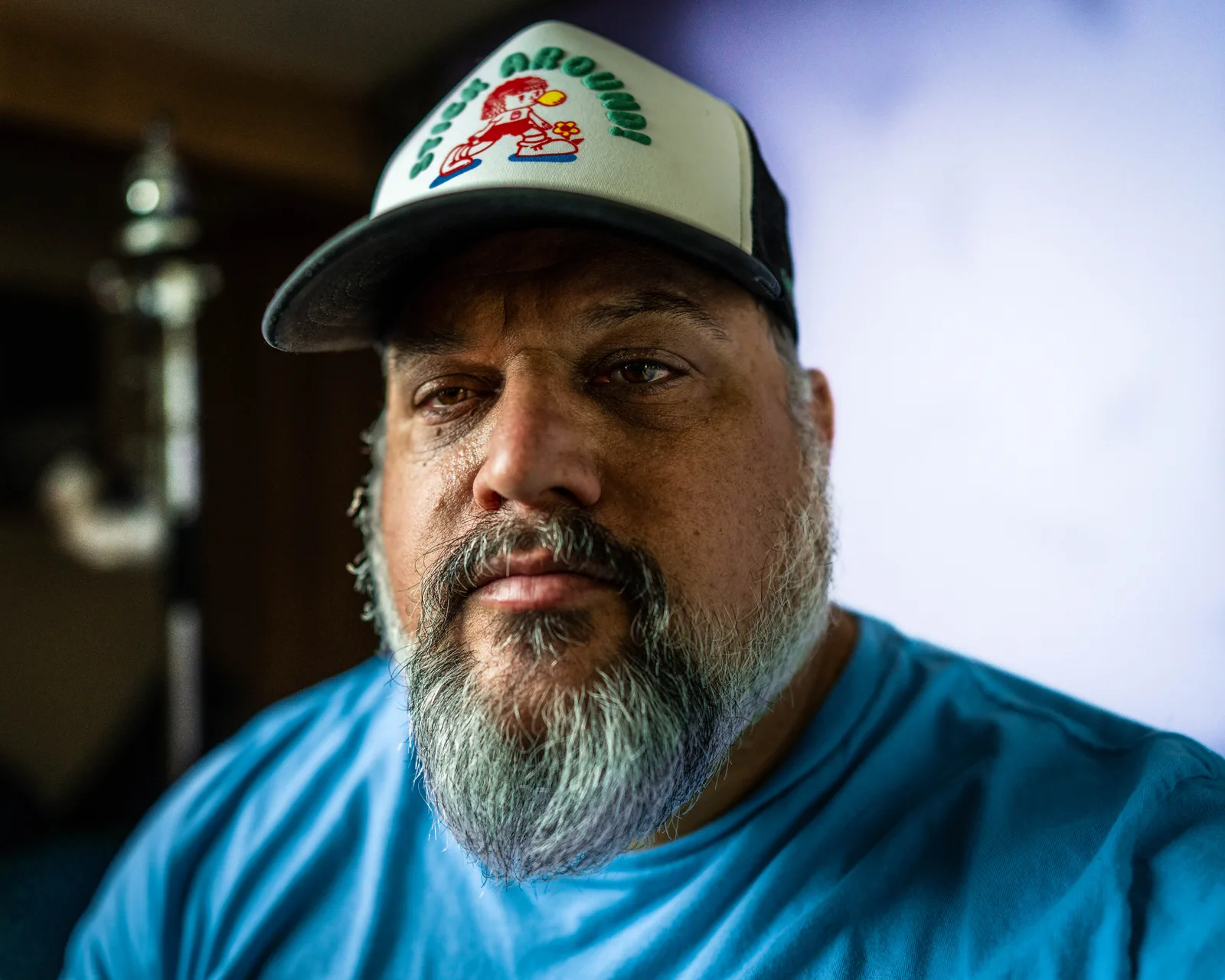 Close-up portrait of a bearded man in a dark cap and blue t-shirt, weathered face and graying beard, looking directly at the camera.