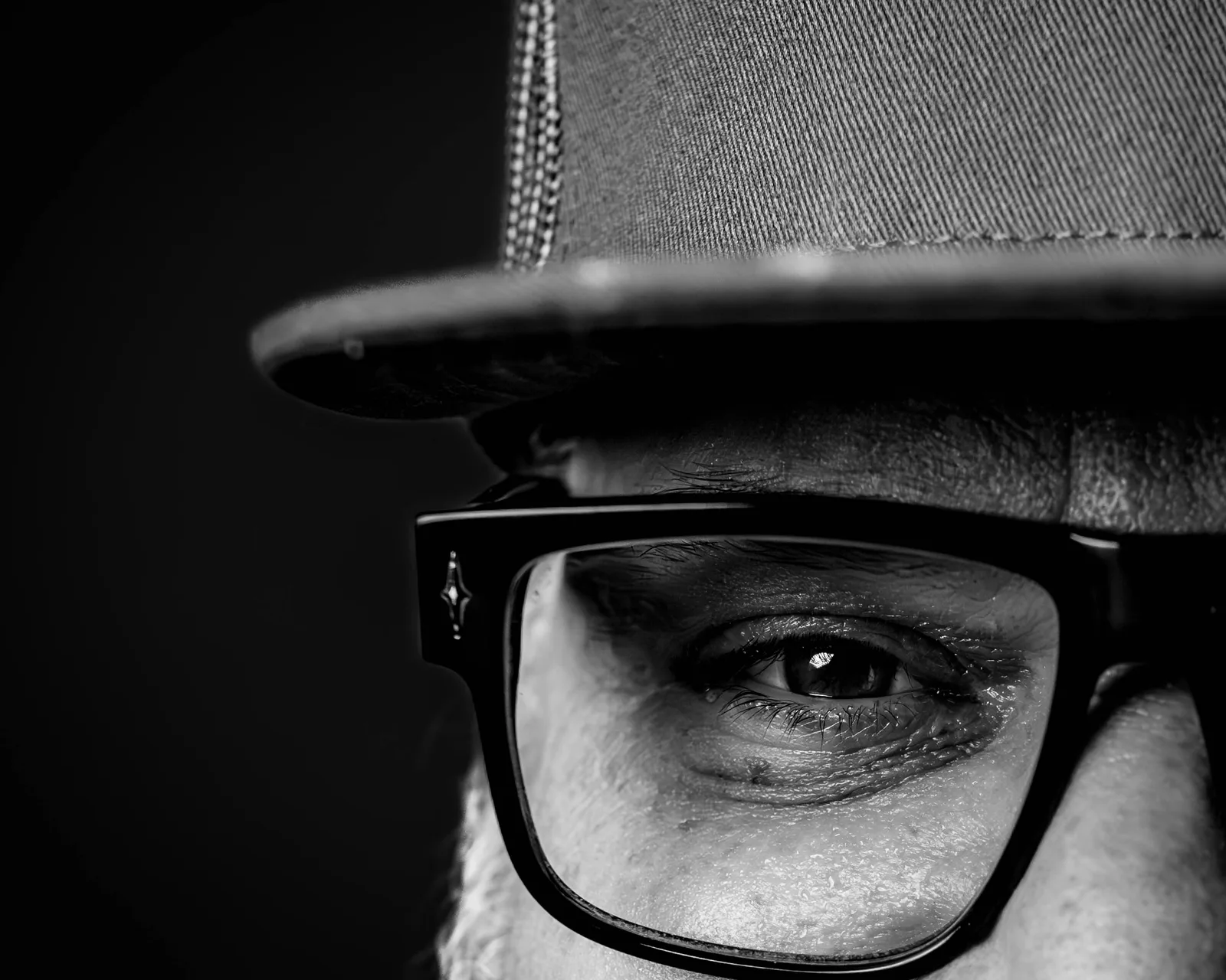 Black-and-white extreme close-up of a man's right eye behind thick-framed glasses, brow furrowed beneath a cap.