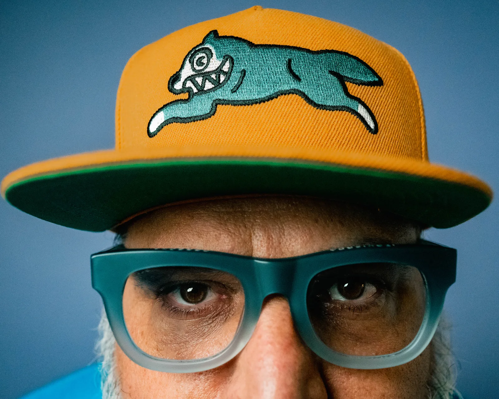 Extreme close-up of a bearded man's eyes behind large blue-gray glasses, under the brim of a yellow and green cap.
