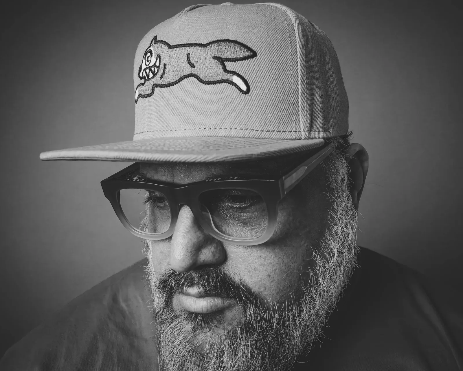 Black-and-white close-up of a bearded man's face under a flat-brim cap with a cartoon running dog patch, wearing thick-framed glasses.