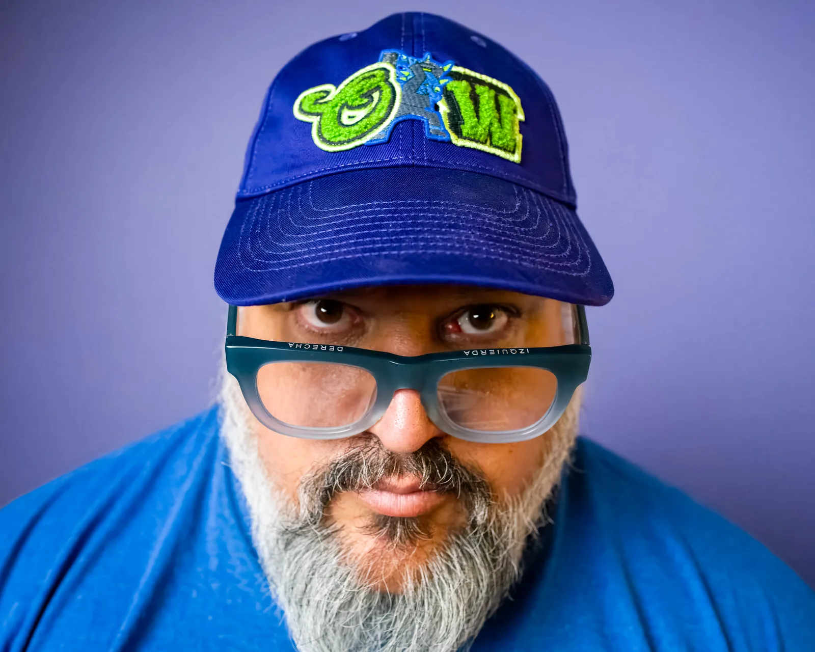 Bearded man in blue cap and shirt stares into camera over slipped-down glasses labeled Izquierda and Derecha, purple backdrop.