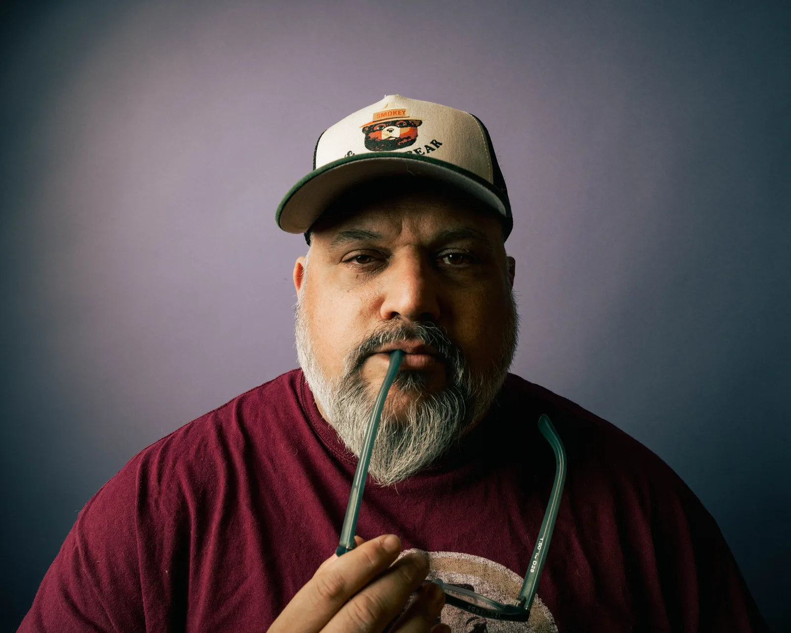 Bearded man in maroon t-shirt and trucker cap holds eyeglasses to his lips, gazing directly at camera against gray backdrop.