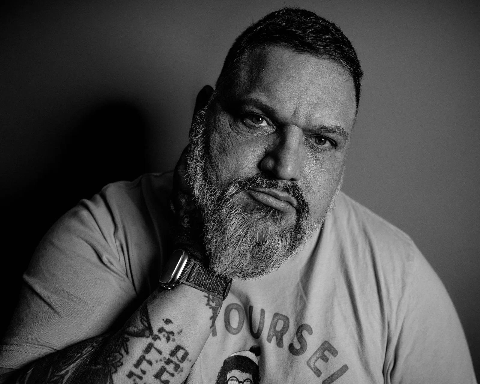 Black-and-white close-up of a bearded man resting his chin on a tattooed hand, wearing a t-shirt reading YOURSELF.