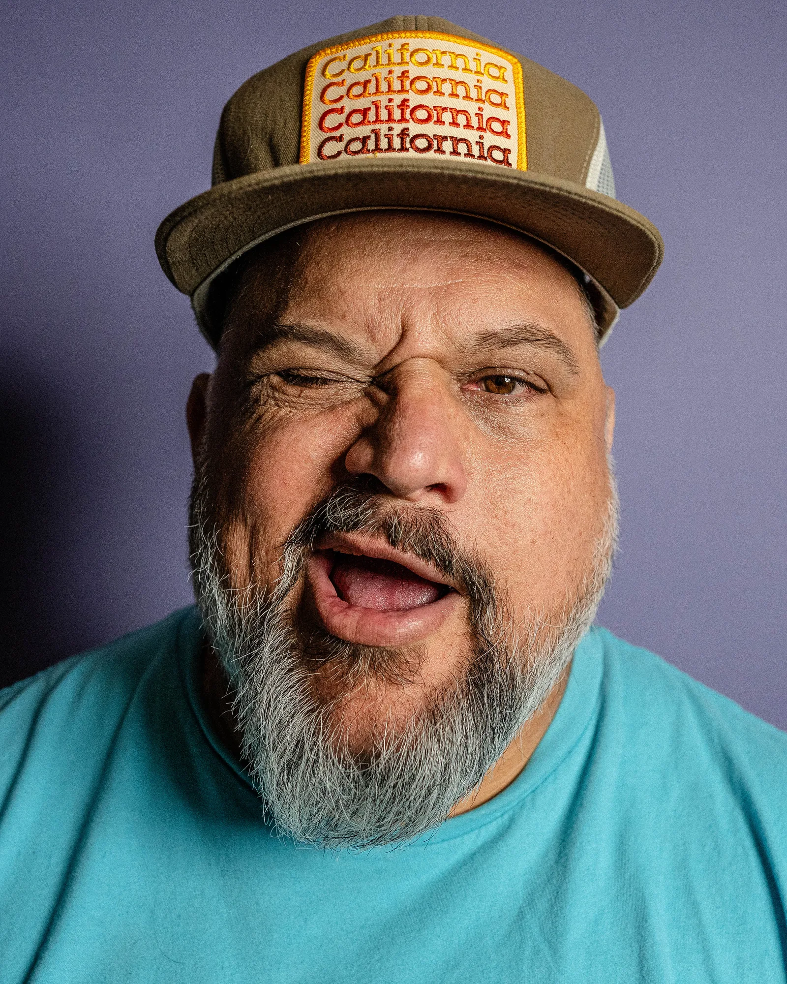 Extreme close-up of a bearded man in a cap and teal shirt, face scrunched mid-expression, mouth open, against a purple backdrop.