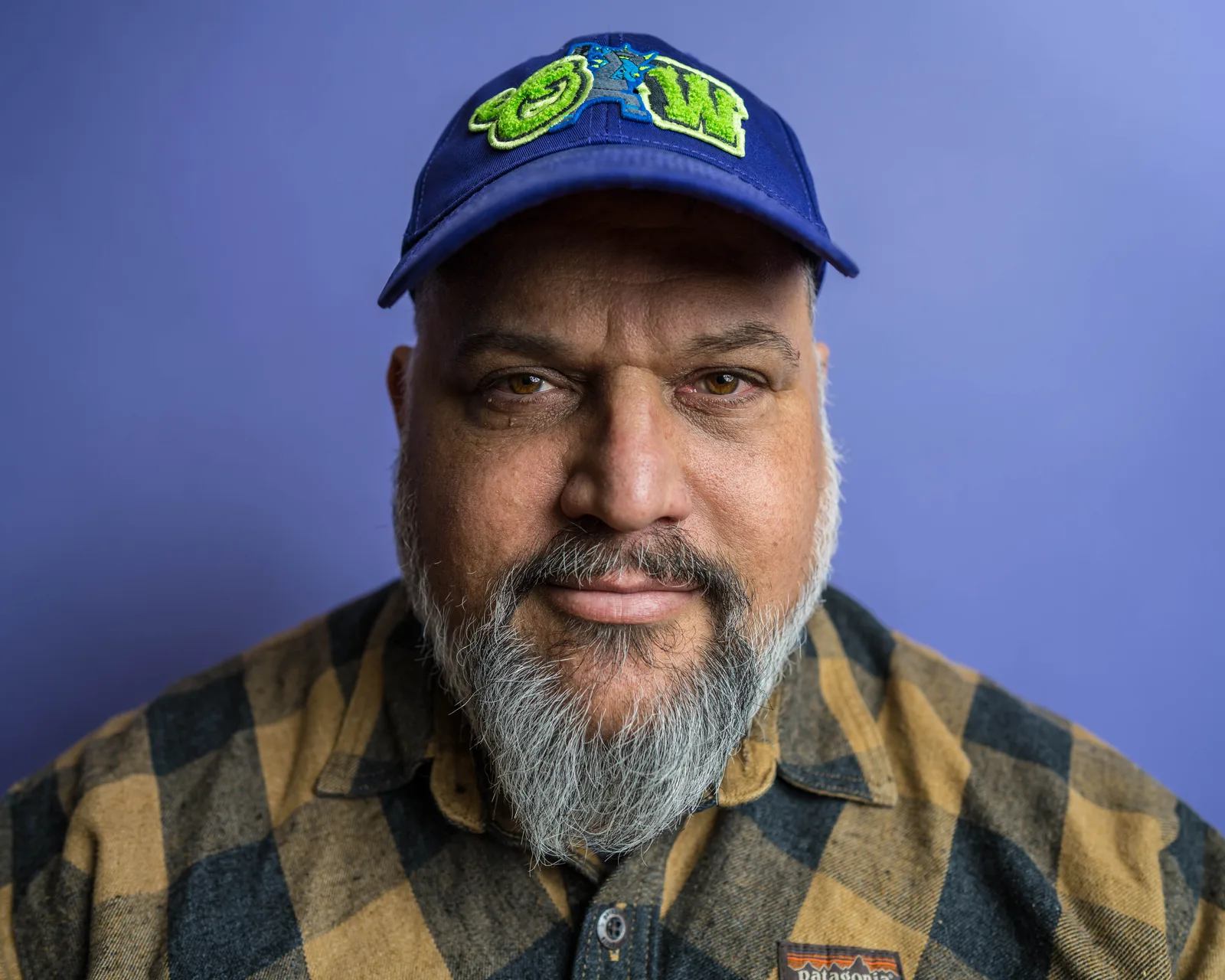 Close-up portrait of a bearded man in a blue cap and plaid flannel shirt against a purple backdrop, gazing directly at the camera.