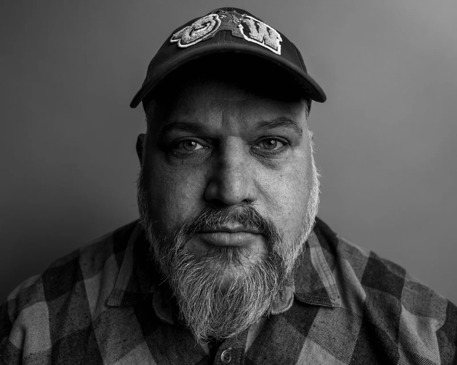 Black-and-white tight portrait of a bearded man in a baseball cap and flannel shirt, eyes glistening, gazing directly at camera.
