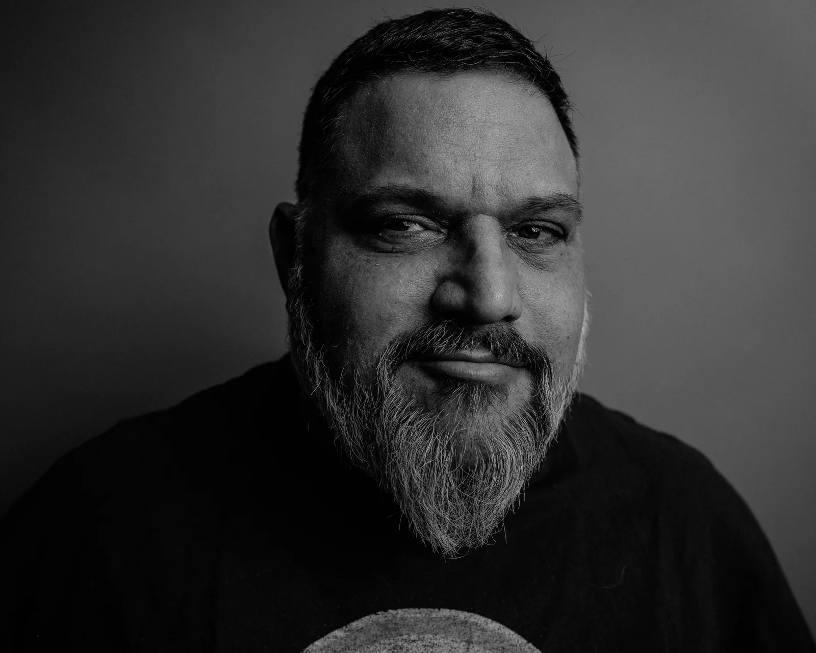 Black-and-white close-up portrait of a bearded man with short hair, faint smile, looking directly at the camera against a gray backdrop.
