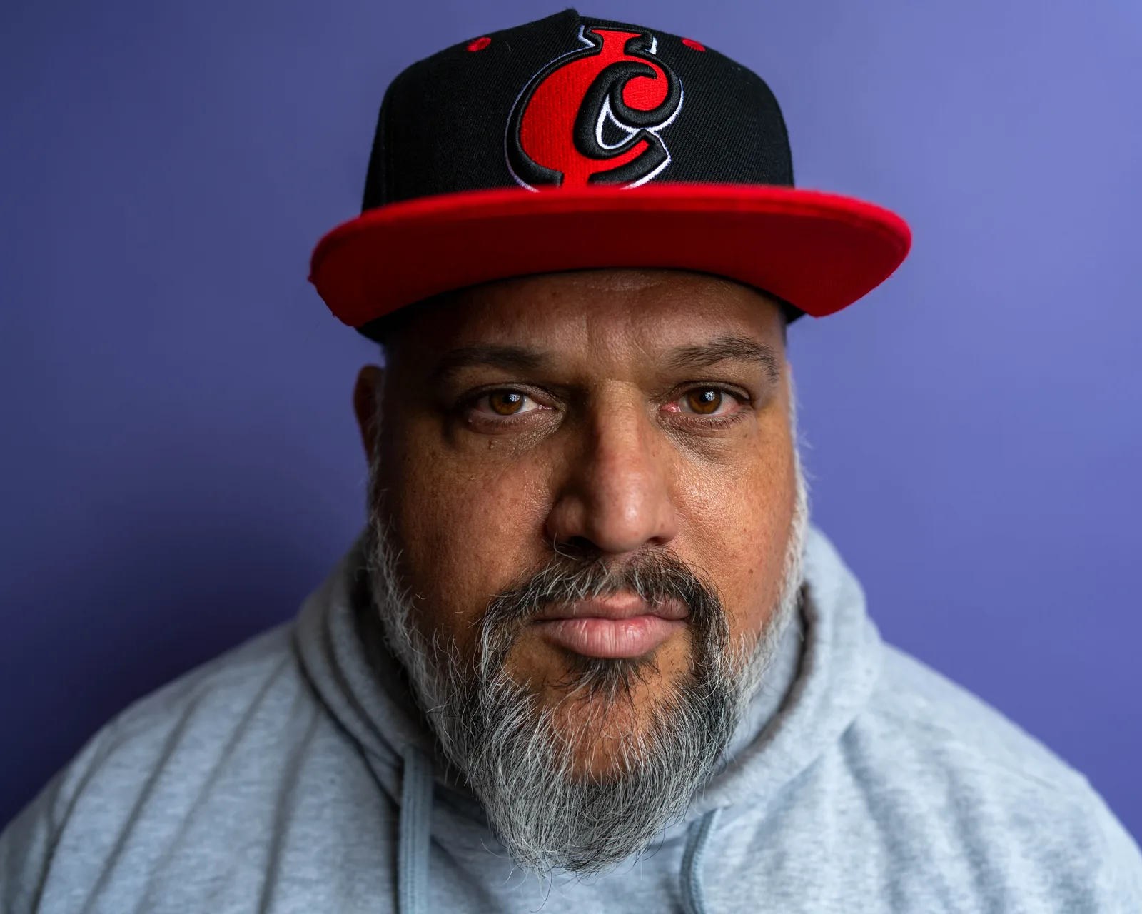 Tight close-up of a bearded man in a gray hoodie and red cap against a purple background, gazing directly at camera.