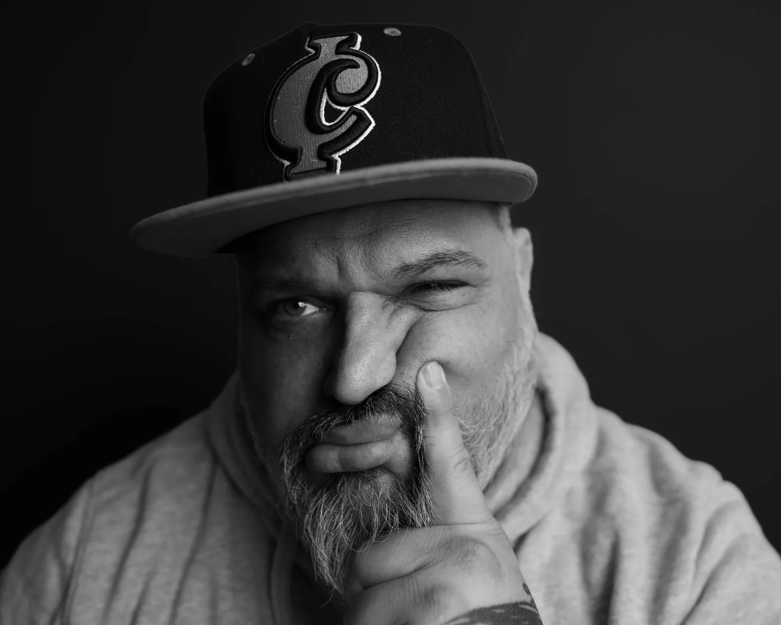 Black-and-white close-up of a bearded man in a hoodie and cap, finger pressed against his upper lip, eyes narrowed at the camera.