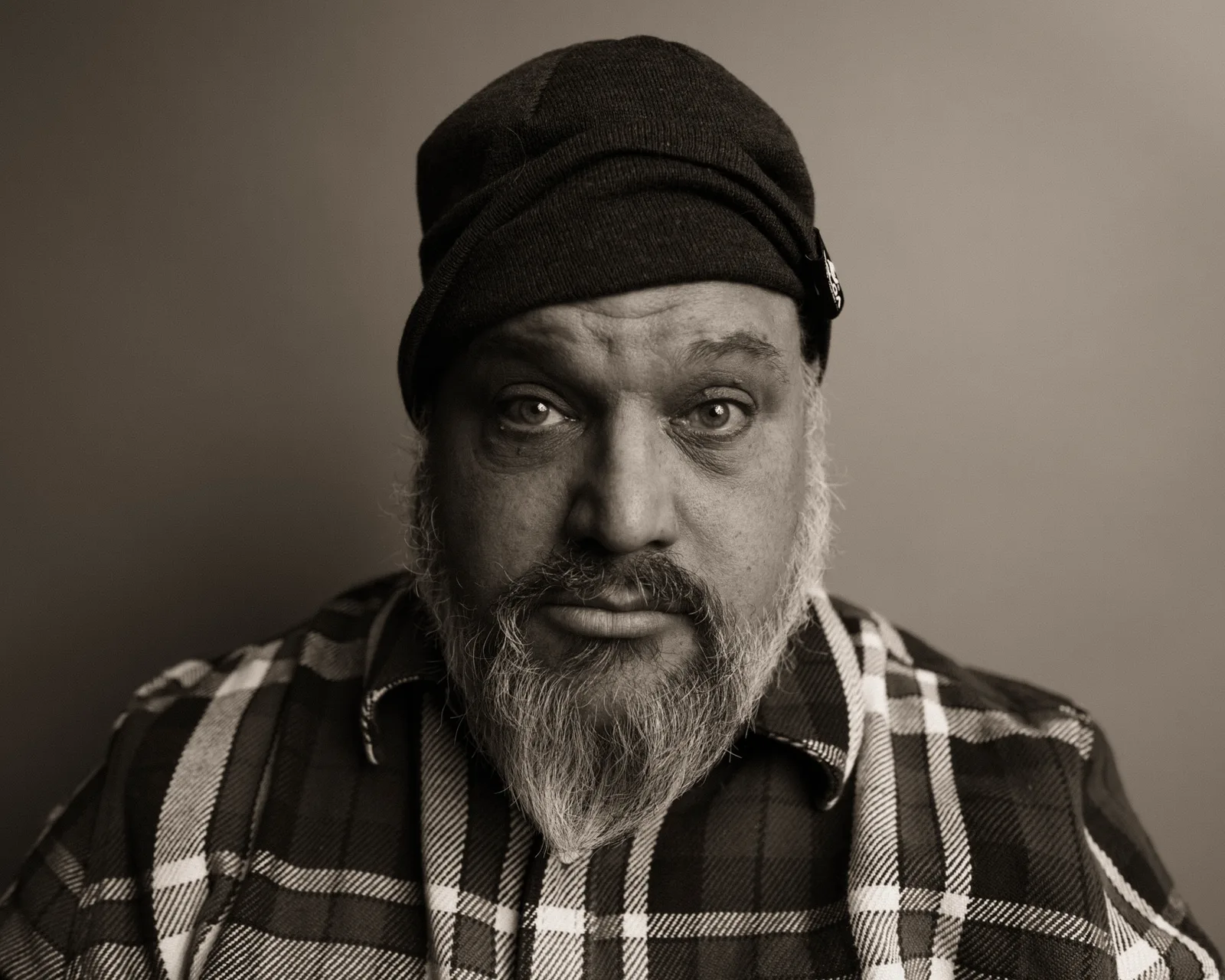 Black-and-white close-up portrait of a bearded man in a plaid flannel shirt, gazing directly at the camera with a steady expression.