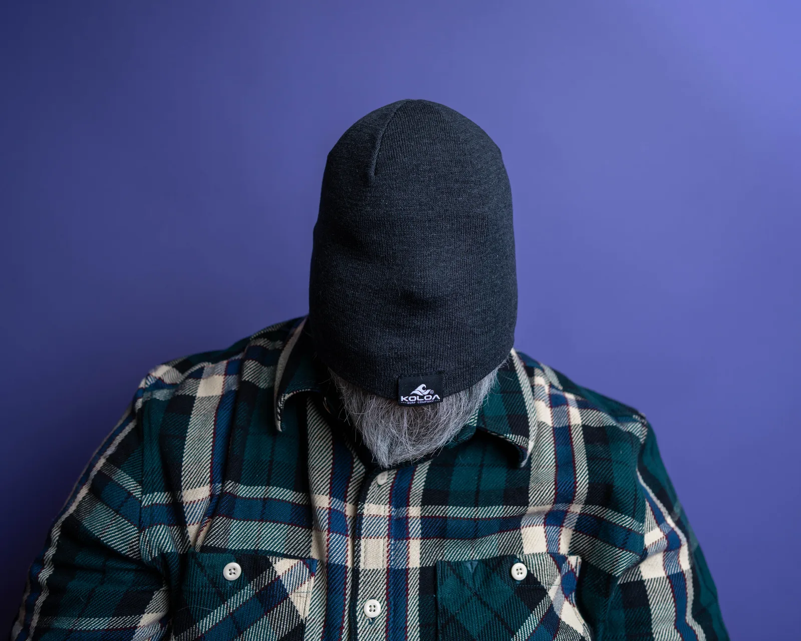 Man in dark beanie pulled low over his face and a green and blue plaid flannel shirt, gray beard visible, against a purple backdrop.