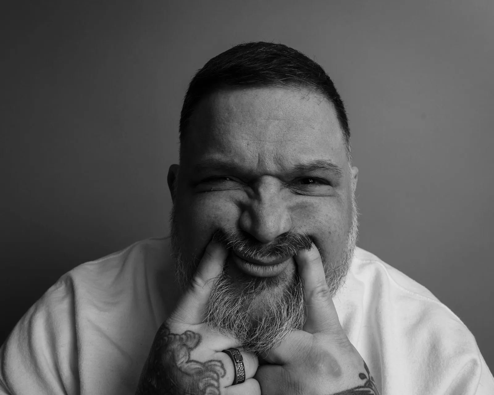 Black-and-white close-up of a bearded man pulling the corners of his mouth wide with tattooed fingers, eyes squinting, in a white shirt.