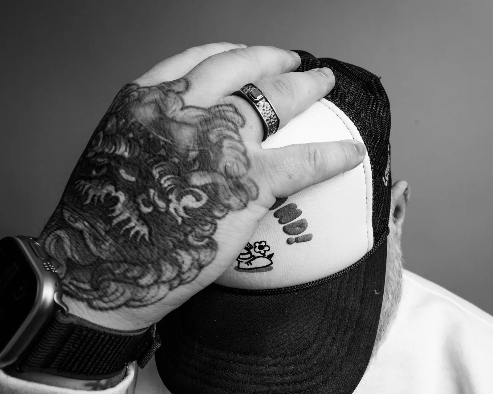 Black-and-white close-up of a heavily tattooed hand with a hammered ring pulling a black trucker hat down over the face