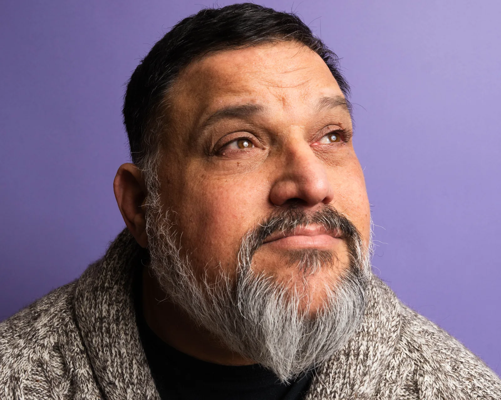 Close-up of a bearded man with graying whiskers looking upward, soft expression, gray knit collar, against a purple backdrop.