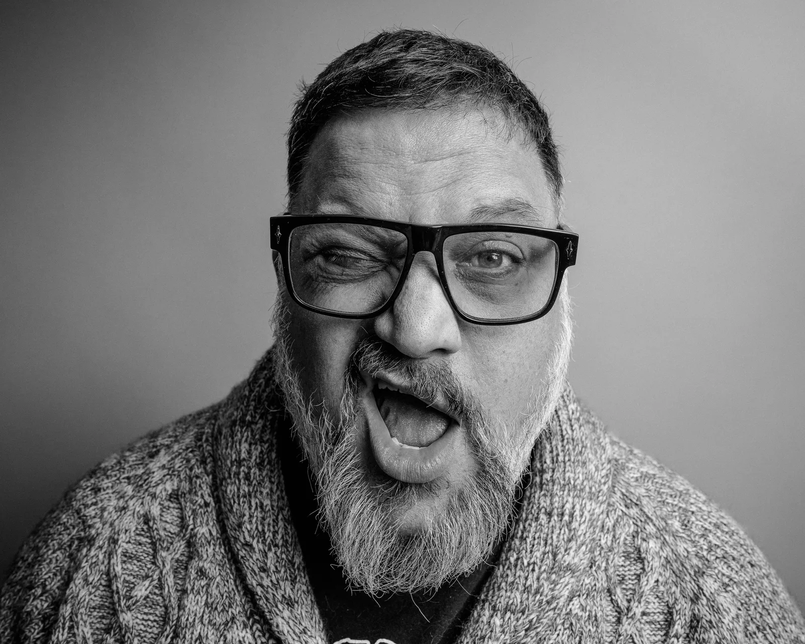 Black-and-white close-up of a bearded man in thick black glasses, tongue out playfully, one eye squinting.