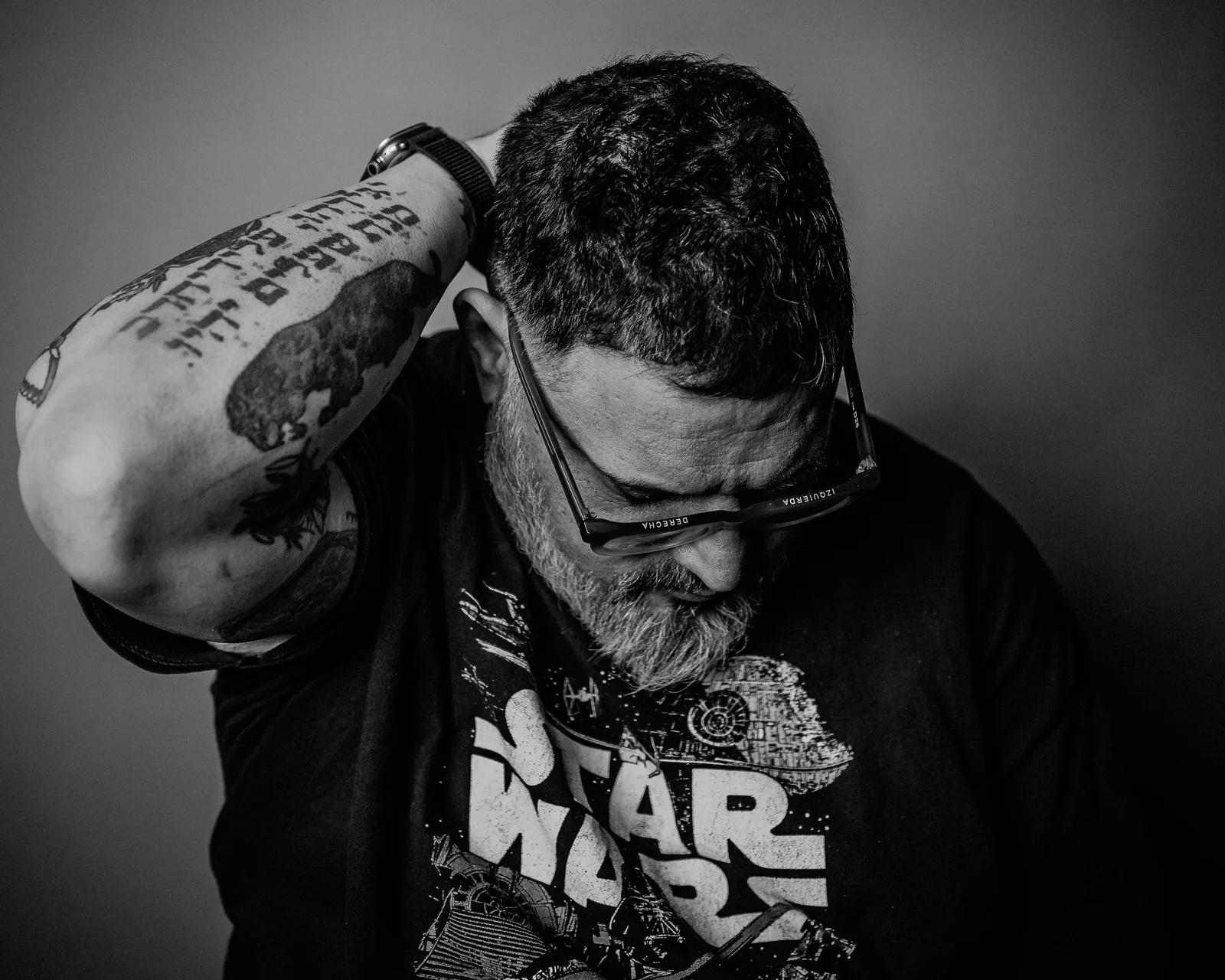 Black-and-white close-up of a bearded man in glasses, head bowed, arm raised behind his head showing heavy tattoos.