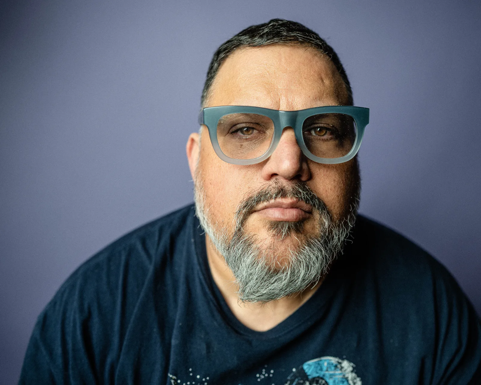 Close-up of a bearded man in oversized teal glasses and a dark t-shirt, looking directly at the camera against a purple-gray backdrop.
