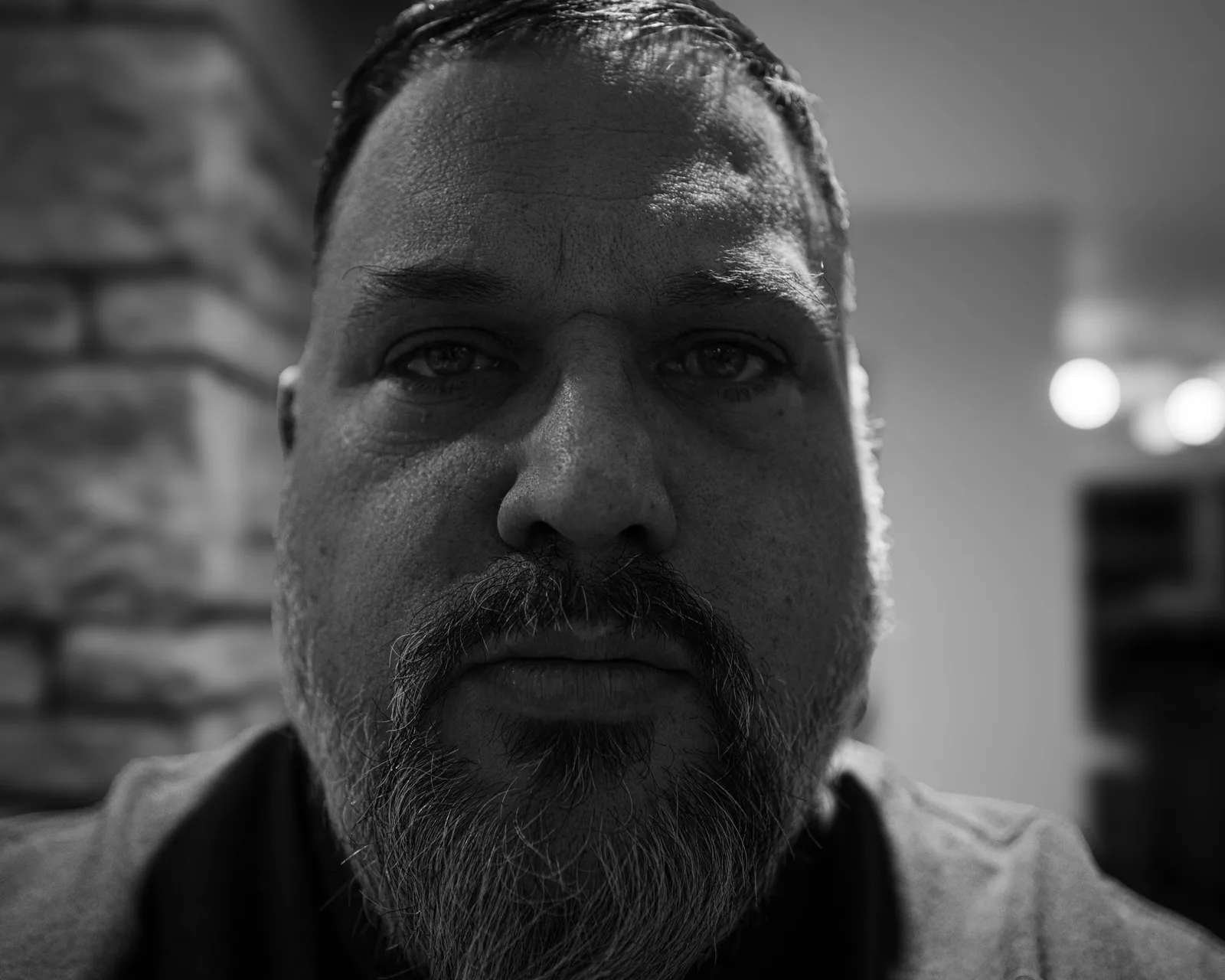 Black-and-white extreme close-up of a man's face, calm direct gaze, mustache and beard, low warm light catching skin