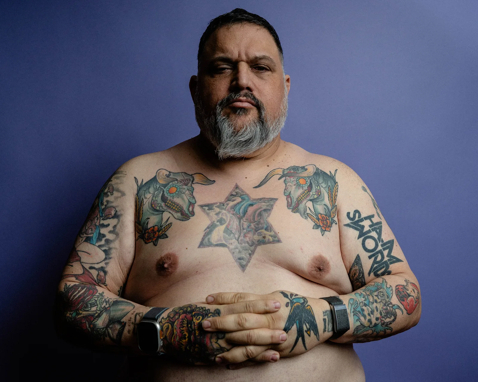 Shirtless torso of a heavily tattooed man, hands clasped at waist, Apple Watch on wrist, gray beard visible at top, blue backdrop.